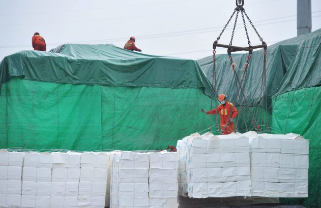 Trabalhadores em uniformes laranja lidam com carga. Um guia fardos brancos sendo içados. Outros estão em cima de pilhas cobertas por lona verde.