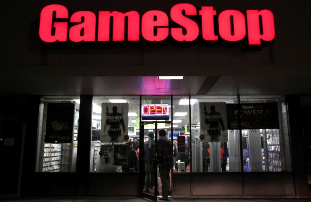Fachada noturna de uma loja GameStop com letreiro vermelho aceso, sinal "OPEN" na porta e pessoas entrando.