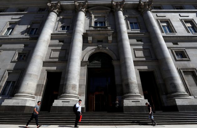 Pessoas caminham em frente a um banco imponente com colunas e escadaria de pedra.