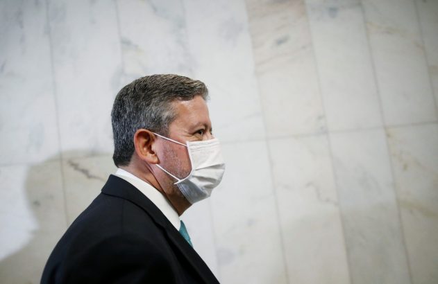 Homem de terno, máscara branca, cabelo grisalho, de perfil, olhando à direita.