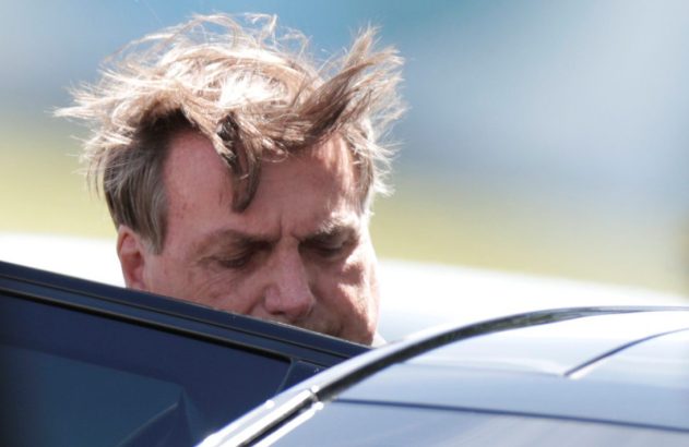 Jair Bolsonaro com cabelo despenteado, olhando para baixo, inclinado sobre um carro escuro.