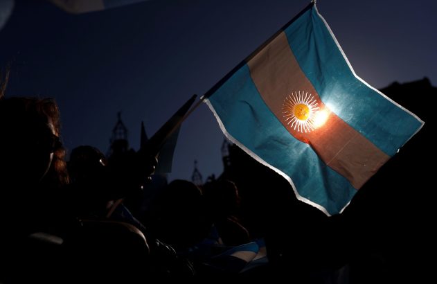 Bandeira da Argentina com Sol de Maio brilhando pela luz; silhuetas de pessoas ao fundo.