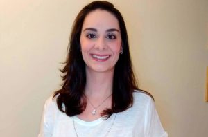 Mulher de cabelos castanhos e olhos escuros sorrindo para a câmera, usando camiseta branca e colar.