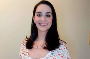 Mulher sorridente de cabelo escuro, com blusa branca estampada com borboletas coloridas e colar.