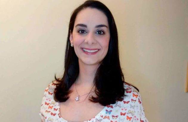 Mulher sorridente de cabelo escuro, com blusa branca estampada com borboletas coloridas e colar.