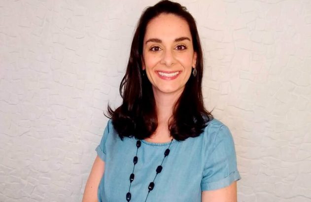 Mulher morena sorri para a câmera, vestindo blusa azul e colar preto, em frente a uma parede branca texturizada.