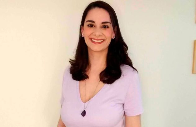 Mulher sorrindo, com cabelos escuros, camiseta lilás e colar com pingente. Fundo claro.