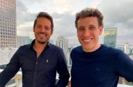 Dois homens sorridentes, olhando para a câmera, em um terraço com paisagem urbana ao fundo.