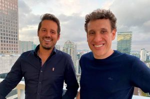 Dois homens sorridentes, olhando para a câmera, em um terraço com paisagem urbana ao fundo.