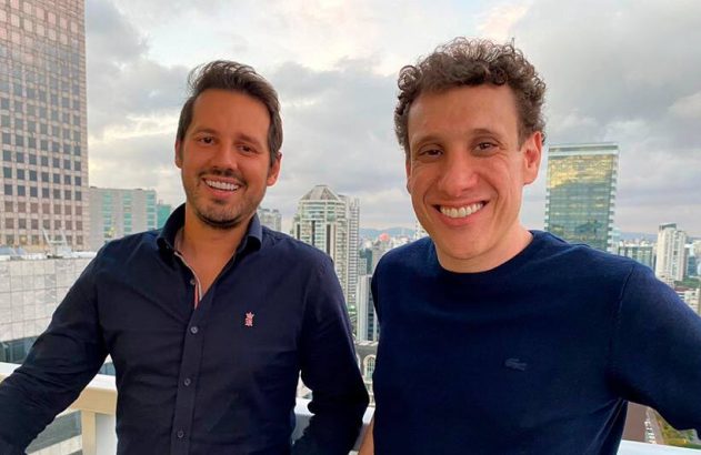 Dois homens sorridentes, olhando para a câmera, em um terraço com paisagem urbana ao fundo.