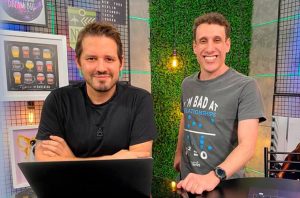 Dois homens em estúdio. Um sentado com laptop, o outro em pé, ambos sorrindo para a câmera.