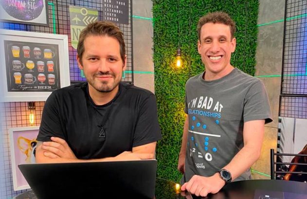 Dois homens em estúdio. Um sentado com laptop, o outro em pé, ambos sorrindo para a câmera.