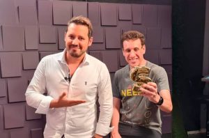 Dois homens sorrindo em um estúdio. Um aponta para o outro, que segura um troféu dourado em forma de S.