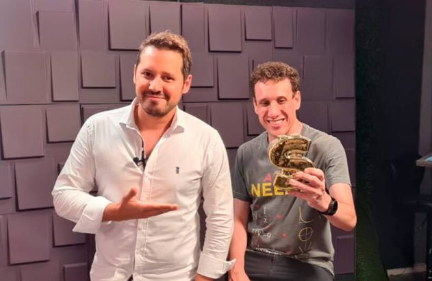 Dois homens sorrindo em um estúdio. Um aponta para o outro, que segura um troféu dourado em forma de S.