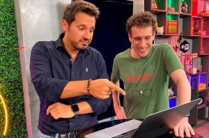 Dois homens olham laptop: um aponta para a tela, o outro sorri. Cenário de estúdio.