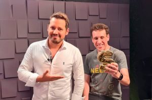 Dois homens sorrindo. Um apresenta o outro, que segura um troféu dourado em forma de S.