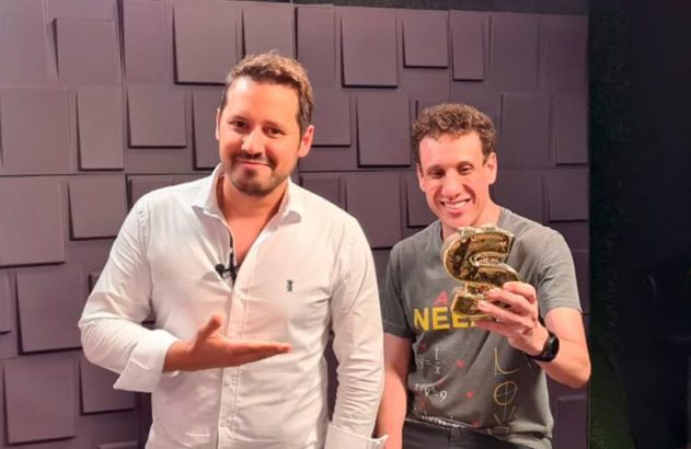 Dois homens sorrindo. Um apresenta o outro, que segura um troféu dourado em forma de S.