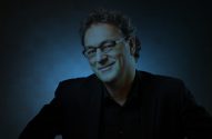 Retrato: homem sorridente de óculos, cabelo escuro, roupa preta, olhando para a câmera, fundo azul.
