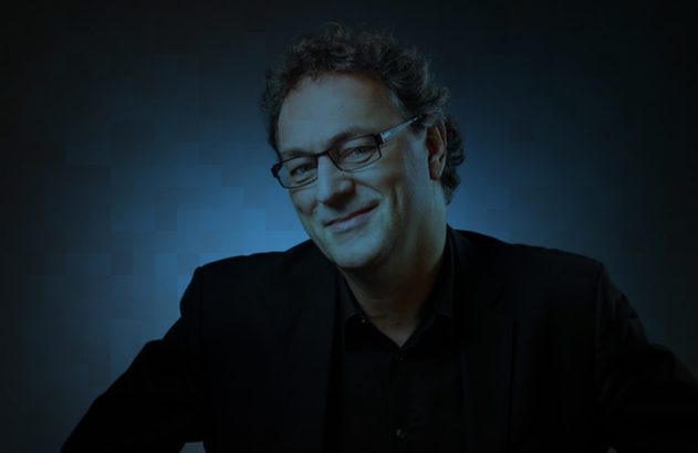 Retrato: homem sorridente de óculos, cabelo escuro, roupa preta, olhando para a câmera, fundo azul.