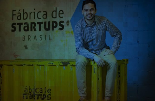 Homem sorridente sentado em contêiner amarelo. Ao fundo, logo Fábrica de Startups Brasil.