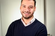 Homem sorridente, suéter azul, camisa branca, braços cruzados, relógio no pulso. Fundo claro.