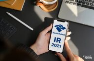 Mãos seguram smartphone exibindo app da Receita Federal com 'IR 2021'. No fundo, laptop e café.