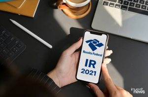 Mãos seguram smartphone exibindo app da Receita Federal com 'IR 2021'. No fundo, laptop e café.