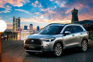 SUV Toyota Corolla Cross prateado em primeiro plano com vista para arranha-céus e céu alaranjado ao pôr do sol.