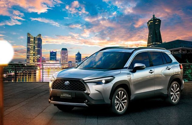 SUV Toyota Corolla Cross prateado em primeiro plano com vista para arranha-céus e céu alaranjado ao pôr do sol.