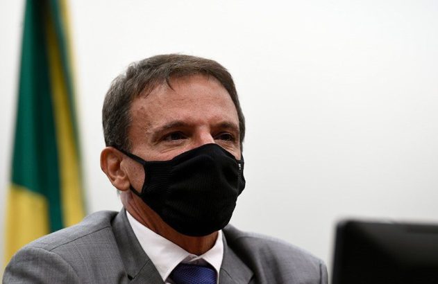 Retrato de homem de terno cinza e máscara preta, olhando para a direita. Fundo branco com bandeira.