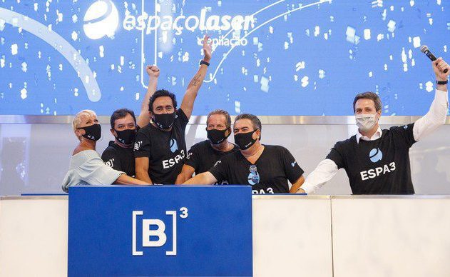 Seis pessoas mascaradas, com braços levantados, celebram atrás de um balcão com o logo B3. Fundo azul e Espaçolaser.