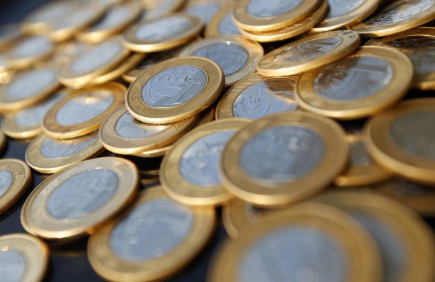 Moedas brasileiras de 1 Real espalhadas sobre uma superfície escura, com algumas em foco nítido.