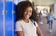 Jovem negra sorrindo, encostada em armários azuis de escola, com alunos desfocados ao fundo.