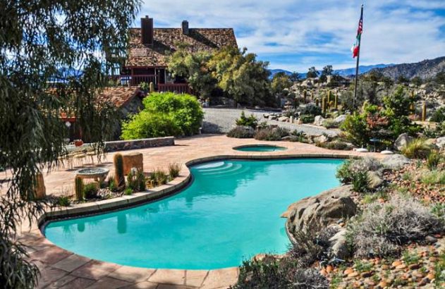 Vista de um resort com telhado de telha, piscina, jacuzzi e paisagismo desértico. Céu claro e montanhas ao fundo.