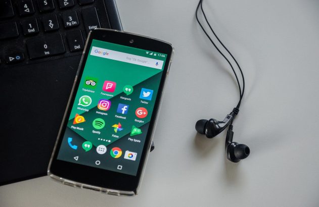 Smartphone com apps na tela, teclado de notebook e fones de ouvido pretos em superfície clara.