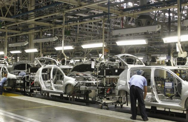 Fábrica de automóveis com carros em montagem na linha de produção e operários trabalhando.