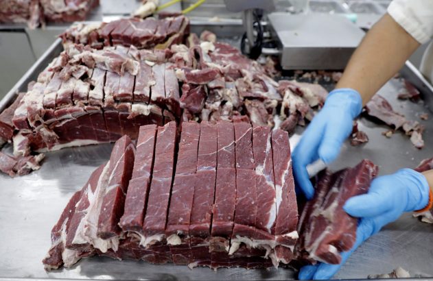 Carne crua fatiada em bancada de metal. Mãos com luvas azuis manuseiam os pedaços de carne.