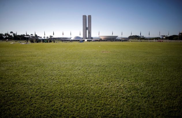 Vista do Congresso Nacional e seu gramado. Um grupo de pessoas está reunido à esquerda, com bandeiras e objetos no chão.
