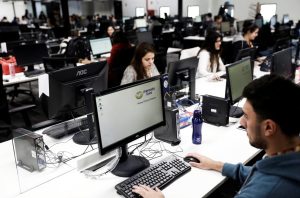 Escritório com várias pessoas trabalhando em computadores. Em primeiro plano, um homem digita e uma tela mostra o logo Mercado Livre.