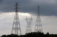 Três imponentes torres de energia com fios e esferas de sinalização contra céu cinzento e nuvens densas sobre árvores.