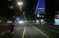 Motociclista entregador em avenida iluminada à noite, com prédios da cidade ao fundo e edifício azul em destaque.