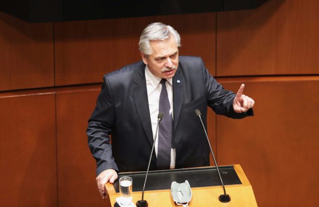Homem de terno discursa em púlpito, gesticulando com o dedo indicador. Há microfones, um copo d'água e uma máscara.