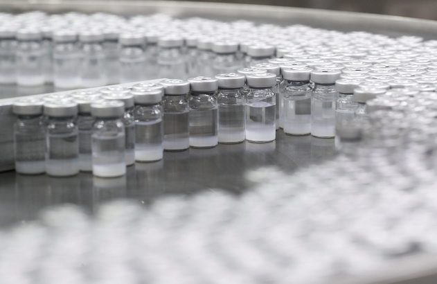 Filas de frascos de vidro com líquido claro e sedimento branco, em linha de produção industrial.