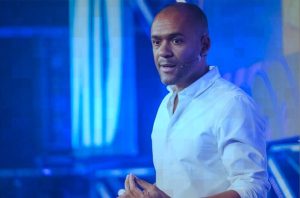 Homem negro em camisa branca, com microfone na orelha, fala em palco com iluminação azul. Mãos unidas.