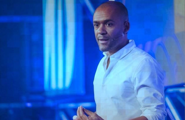 Homem negro em camisa branca, com microfone na orelha, fala em palco com iluminação azul. Mãos unidas.