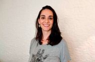 Uma mulher de cabelo escuro e camiseta cinza com estampa de girafa sorri para a câmera. Fundo branco texturizado.
