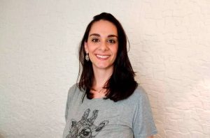 Uma mulher de cabelo escuro e camiseta cinza com estampa de girafa sorri para a câmera. Fundo branco texturizado.