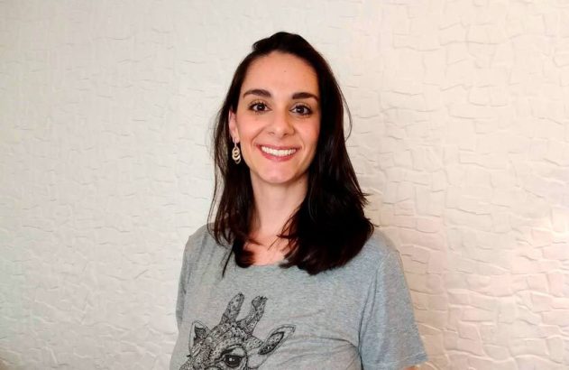 Uma mulher de cabelo escuro e camiseta cinza com estampa de girafa sorri para a câmera. Fundo branco texturizado.