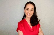 Mulher sorrindo de blusa vermelha e cabelo escuro, braços cruzados, em frente a parede texturizada.