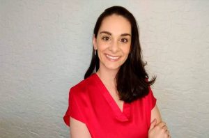 Mulher sorrindo de blusa vermelha e cabelo escuro, braços cruzados, em frente a parede texturizada.